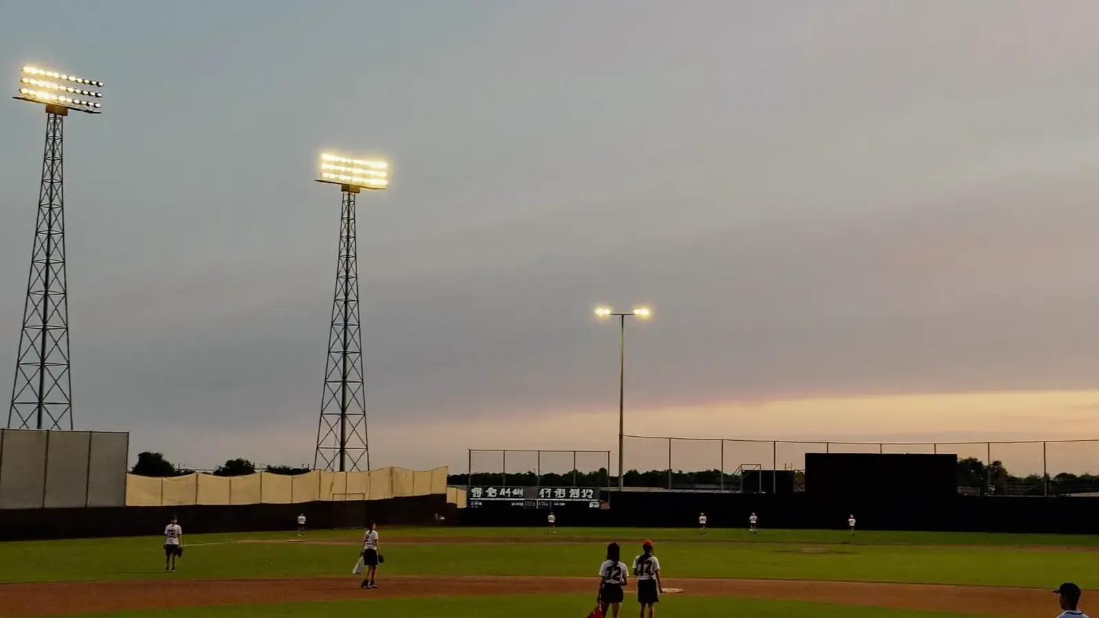 A youth baseball complex at dusk with stadium lights glowing over the field